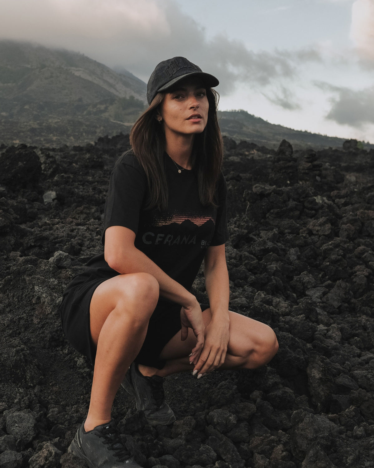 Girl cerana Tshirt and Cap sitting on a rocky landscape with mountains in the background