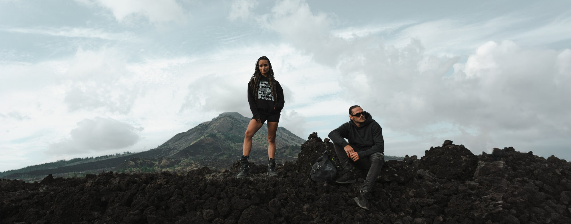 Cerana black hoodie outfit and duffle bag shot on dramatic volcanic terrain with mountain view – urban outdoor aesthetic